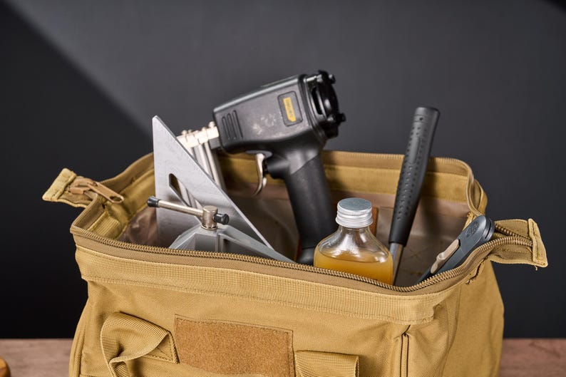 May include: A tan canvas tool bag filled with various tools. The tools include a black and gray nail gun, a silver metal square, a bottle of liquid, a black hammer, and a small gray tool. The bag is open and ready for use.