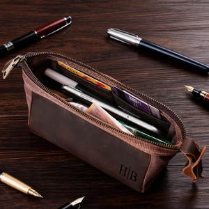 Può includere: Un astuccio marrone in tela e pelle con cerniera, aperto per rivelare penne, un telefono e carte. L'astuccio ha le iniziali "HB" sul lato. Diverse penne sono sparse su una superficie di legno.