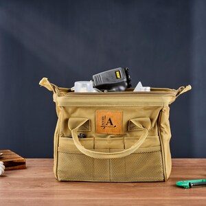 May include: A tan canvas tool bag with multiple pockets and a handle. The bag is open, revealing tools inside. A leather patch with the letter "A" is on the front. The bag is on a wooden surface.