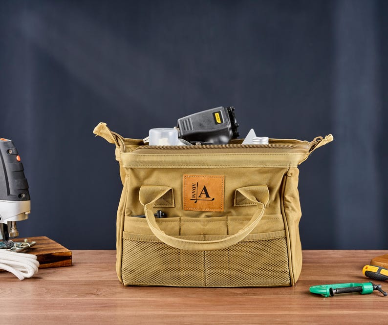 May include: A tan canvas tool bag with multiple pockets and a handle. The bag is open, revealing tools inside. A leather patch with the letter "A" is on the front. The bag is on a wooden surface.