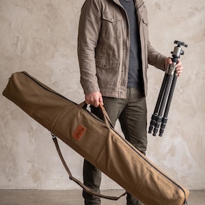May include: A long, tan canvas carrying bag with a black zip, a brown leather handle, and a rectangular brown leather patch featuring the text Helby. The bag includes an adjustable shoulder strap and is designed for transporting equipment.