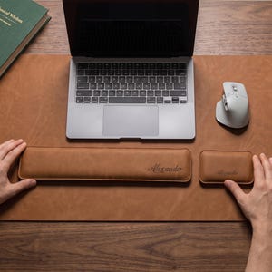 May include: A tan leather desk mat featuring a matching long rectangular keyboard wrist rest and a smaller rectangular mouse wrist rest, both embossed with the name Alexander in a cursive script, positioned below a silver laptop and a white mouse.