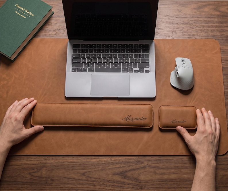 May include: A tan leather desk mat featuring a matching long rectangular keyboard wrist rest and a smaller rectangular mouse wrist rest, both embossed with the name Alexander in a cursive script, positioned below a silver laptop and a white mouse.