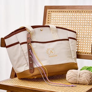 May include: A cream and tan canvas tote bag with brown trim, featuring a vertical leather strip with metal grommets for yarn, two top handles, and a gold snap closure. The front displays a circular logo with the letter A and the name Ameenah.
