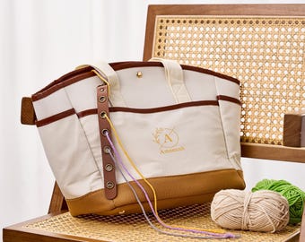 May include: A cream and tan canvas tote bag with brown trim, featuring a vertical leather strip with metal grommets for yarn, two top handles, and a gold snap closure. The front displays a circular logo with the letter A and the name Ameenah.