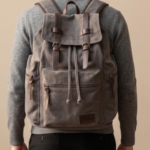 May include: A grey canvas backpack featuring dark brown leather straps with metal buckles, a drawstring closure, and two side pockets with zipper pulls. A rectangular leather patch with embossed text is located on the lower front pocket.