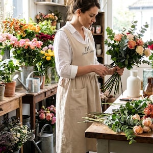 Może przedstawiać: Kwiaciarka w beżowym fartuchu z napisem "JH flower shop", układająca bukiet róż i eukaliptusa. Sklep prezentuje różne kwiaty w donicach i wazonach, z konewkami i drewnianymi stołami.
