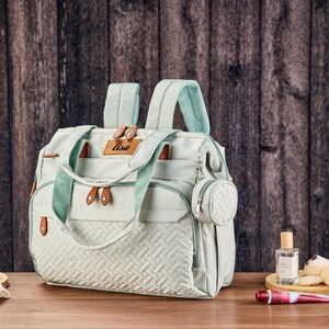 Może przedstawiać: Jasnozielona torba na pieluchy z pikowanym panelem przednim i brązowymi sk&oacute;rzanymi detalami. Torba ma g&oacute;rny uchwyt, paski na ramiona i małą, okrągłą, dołączoną torebkę. Słowo "Lisa" znajduje się na sk&oacute;rzanej metce. Inne przedmioty znajdują się na drewnianej powierzchni.