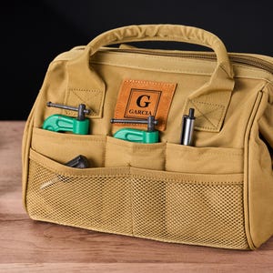 May include: Tan canvas tool bag with multiple pockets, a top handle, and a zipper closure. The bag has a leather patch with the name "GARCIA" and a large "G". It contains green clamps and a pen.
