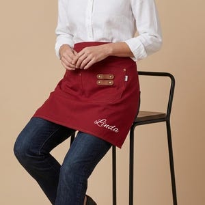 May include: A deep red waist apron featuring two front pockets with brown leather accents secured by brass rivets. The apron displays the name Linda embroidered in white cursive script on the lower right side, providing a functional design.