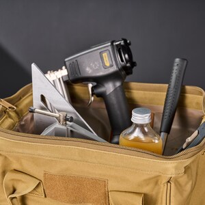 May include: A tan canvas tool bag filled with various tools. The tools include a black and gray nail gun, a silver and gray angle square, a bottle of liquid, a black hammer, and a small gray tool.