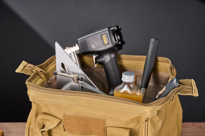 May include: A tan canvas tool bag filled with various tools. The tools include a black and gray nail gun, a silver and gray angle square, a bottle of liquid, a black hammer, and a small gray tool.