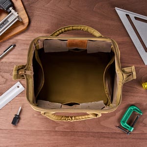 May include: Open tan canvas tool bag with multiple interior pockets and a leather accent. Various tools surround the bag, including a speed square, hammer, and clamp, all arranged on a wooden surface.