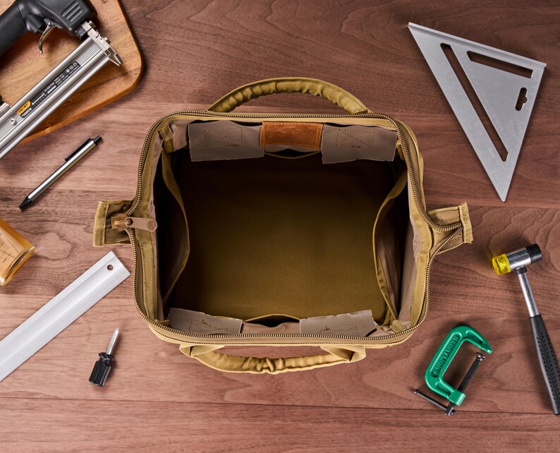 May include: Open tan canvas tool bag with multiple interior pockets and a leather accent. Various tools surround the bag, including a speed square, hammer, and clamp, all arranged on a wooden surface.
