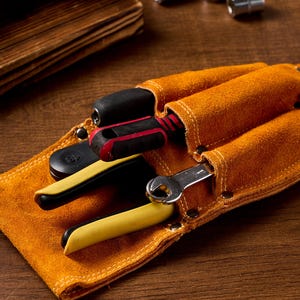 May include: A tan leather tool belt with three pockets, holding a screwdriver, pliers, and a wrench. The tools have black and yellow handles. The belt is on a wooden surface.