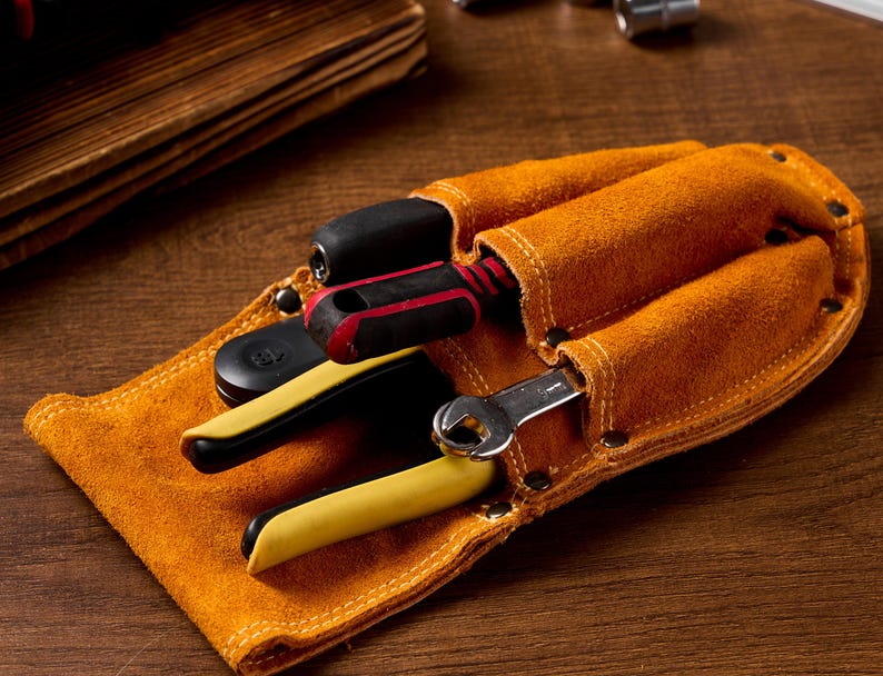 May include: A tan leather tool belt with three pockets, holding a screwdriver, pliers, and a wrench. The tools have black and yellow handles. The belt is on a wooden surface.
