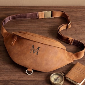 May include: A tan leather fanny pack featuring a dark brown monogram with the name MARLON and the letter M. The bag includes an adjustable brown strap with a metallic buckle, a front zipper, and a silver metal D-ring at the bottom base.