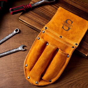 May include: A tan leather tool belt with three pockets and the name "SEAN" in a decorative font. Two spanners and a ratchet are also visible. The tools are on a wooden surface.