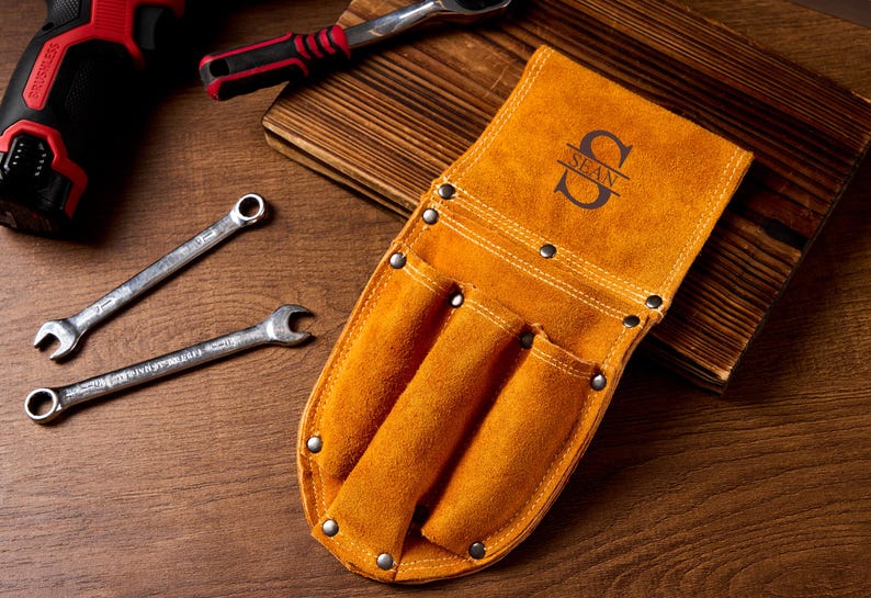 May include: A tan leather tool belt with three pockets and the name "SEAN" in a decorative font. Two spanners and a ratchet are also visible. The tools are on a wooden surface.