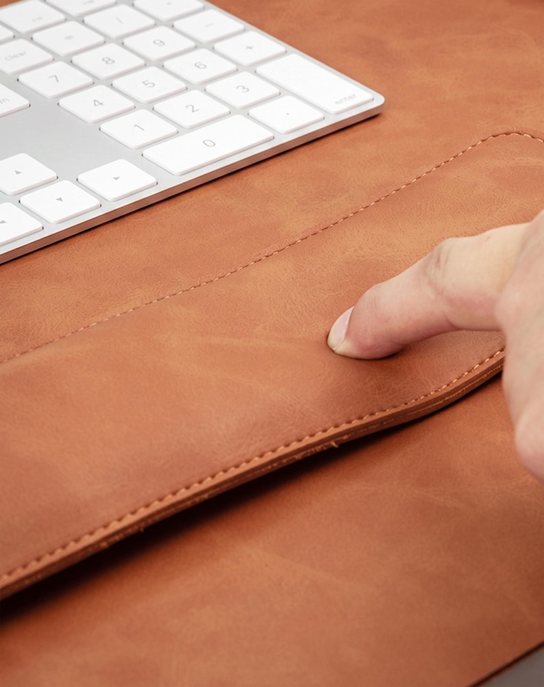May include: A tan leather desk mat with a smooth, matte surface and stitched edges, positioned next to a white wireless computer keyboard with a numeric keypad. The material shows a natural grain texture and a soft, flexible appearance.
