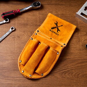 May include: An orange leather tool belt with three pockets, the word "MAXWELL" and a wrench logo. Several spanners, a ratchet, and a metal square are arranged on a wooden surface.