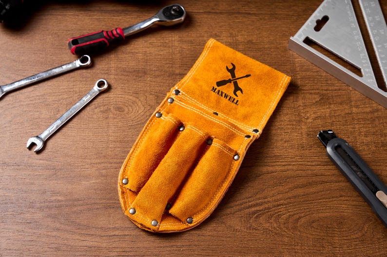 May include: An orange leather tool belt with three pockets, the word "MAXWELL" and a wrench logo. Several spanners, a ratchet, and a metal square are arranged on a wooden surface.