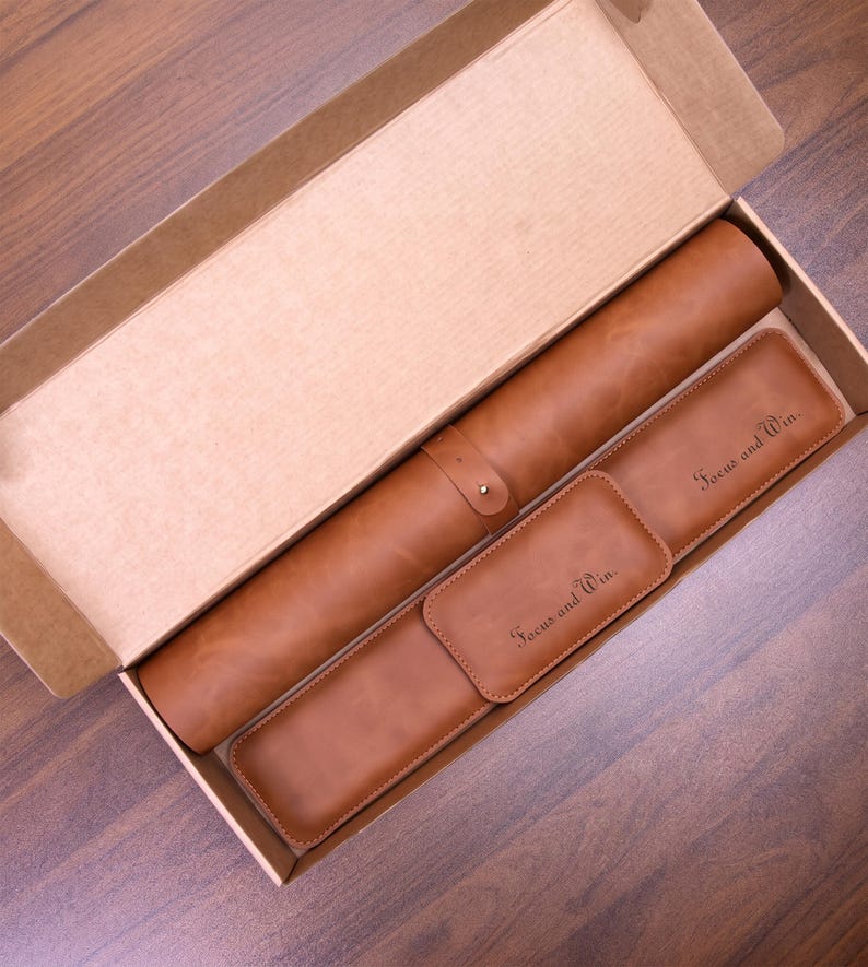 May include: A tan leather set inside a cardboard box, including a cylindrical roll with a strap, a rectangular case, and a small rectangular tag. The case and tag are both embossed with the text Focus and Win in a dark script font.