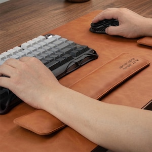 May include: A tan leather desk mat with a matching rectangular wrist rest featuring the coordinates 40&deg;42'46" N 74&deg;00'22" W embossed on the surface. The setup includes a mechanical keyboard with a gradient of white to black keys.
