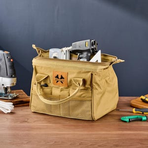 May include: A tan canvas tool bag with a zipper closure, handles, and multiple pockets. The bag contains tools, including a nail gun and a white triangle. A jigsaw and screwdrivers are also visible.