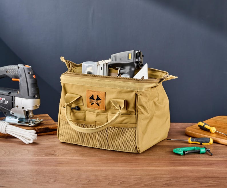 May include: A tan canvas tool bag with a zipper closure, handles, and multiple pockets. The bag contains tools, including a nail gun and a white triangle. A jigsaw and screwdrivers are also visible.