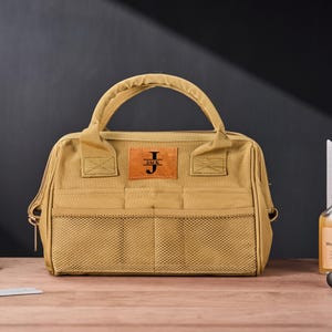 May include: Tan canvas tool bag with a brown leather patch that reads "JACK" with the initial "J". The bag has a handle, mesh pockets, and a zipper closure. A bottle of wood wax oil and a metal square are in the background.