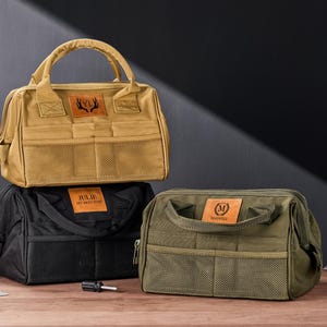 May include: Three canvas tool bags in tan, black, and olive green, each with a handle and multiple pockets. The tan bag has a deer antler logo, the black bag says "JULIE MY BEST SHOP", and the green bag has an "M" logo.