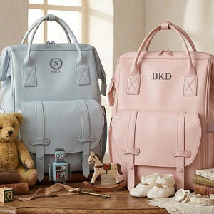 May include: Two leather diaper bags, one light blue with "SMITH" and one pink with "BKD" embossed. Each bag has a top handle, front flap, and side pockets. Accessories include a teddy bear, toy robot, wooden blocks, and baby shoes.