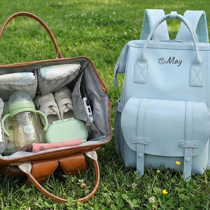 以下が含まれることがあります： 茶色の革製おむつバッグ。上部が開いており、おむつ、哺乳瓶、その他のベビー用品が見えます。 「May」という名前のライトブルーのバックパック型おむつバッグも表示されています。 どちらのバッグも草の上にあります。