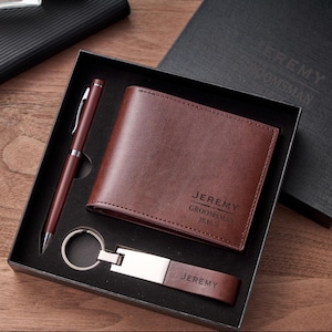 Puede incluir: Un juego de cartera, bolígrafo y llavero de cuero marrón en una caja de regalo negra. La cartera y el llavero están grabados con el nombre "JEREMY". El bolígrafo es de color marrón a juego. La caja está abierta, mostrando el contenido.