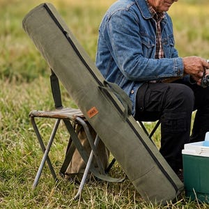 Può includere: Una borsa per canne da pesca in tela verde oliva con una lunga cerniera nera, due manici in tessuto e una piccola toppa rettangolare in pelle marrone con il testo ALEX in rilievo sul pannello laterale esterno.