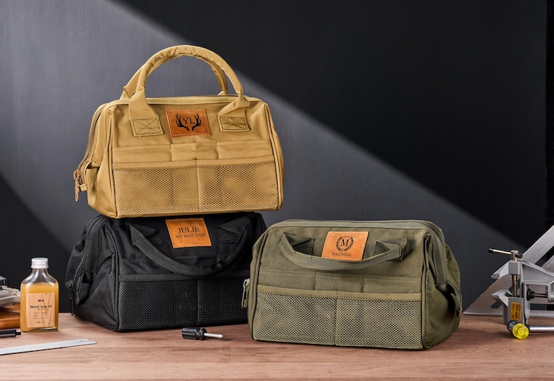 May include: Three canvas tool bags in tan, black, and olive green, each with a top handle, front pockets, and a leather patch. The tan bag has a deer antler logo, the black bag says "JULIE MY BEST SHOP", and the green bag has an "M" logo.