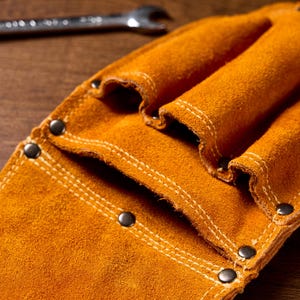 May include: Close-up of a tan leather tool belt with multiple pockets and riveted accents. The tool belt is made of suede leather and has white stitching. A spanner is visible in the background.