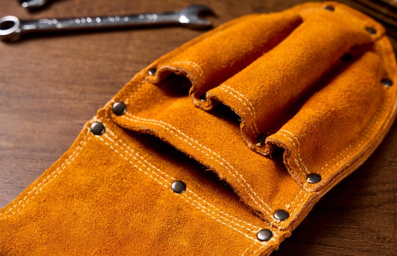 May include: Close-up of a tan leather tool belt with multiple pockets and riveted accents. The tool belt is made of suede leather and has white stitching. A spanner is visible in the background.
