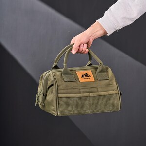 May include: Olive green tote bag with a rectangular shape, featuring a top handle and a front mesh pocket. A brown leather patch with a mountain logo and the word "SOURCE" is visible. The bag is held by a person.