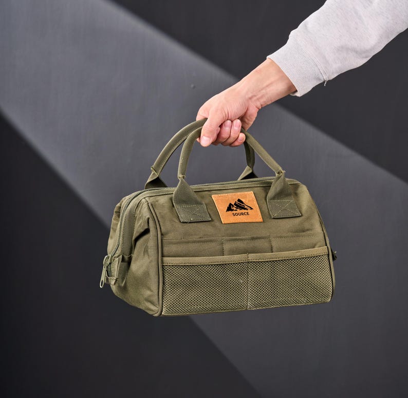 May include: Olive green tote bag with a rectangular shape, featuring a top handle and a front mesh pocket. A brown leather patch with a mountain logo and the word "SOURCE" is visible. The bag is held by a person.