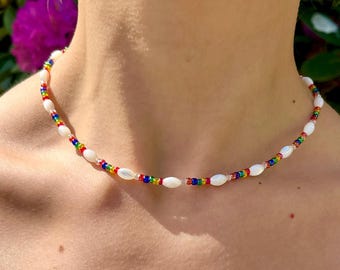 May include: A beaded necklace featuring white oval beads interspersed with small rainbow-colored beads. The necklace is displayed against a natural background, highlighting its colorful and delicate design. This necklace is a fashion accessory.