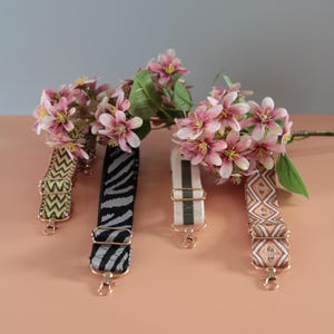 Peut inclure: Quatre bandoulières de sac à main décoratives avec des fermoirs dorés, chacune avec un motif unique : zigzag, zébré, rayé et géométrique. Des fleurs roses et des feuilles vertes sont en arrière-plan.