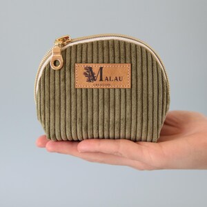 May include: Olive green corduroy pouch with a gold zipper and a leather patch that reads "MALAU CREATION". The pouch is held in a hand against a light gray background. The pouch is small and round.
