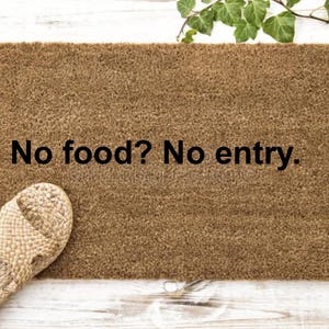 Può includere: Un zerbino rettangolare marrone con la scritta "No food? No entry." è in mostra. Sandali intrecciati sono a sinistra. L'edera verde è nell'angolo in alto a destra. Lo zerbino è su una superficie di legno bianco.