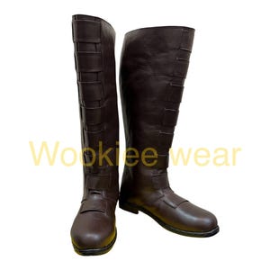 Puede incluir: Un par de botas marrones hasta la rodilla. Las botas tienen un aspecto de cuero liso con correas y hebillas decorativas. Las botas están diseñadas para usar y están sobre un fondo blanco. El texto "Wookiee wear" es visible.