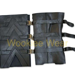 May include: Black leather arm guards with buckle fastenings and decorative stitching. The arm guards feature a geometric design and are secured with adjustable straps. The text "Wookiee Wear" is visible.