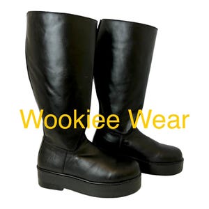 May include: A pair of black knee-high boots with a thick platform sole and a smooth finish. The text Wookiee Wear is displayed in yellow across the center of the boots. The boots feature a rounded toe and a tall shaft that reaches the lower leg.