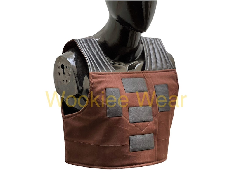 May include: Brown vest with black detailing, displayed on a black mannequin. The vest has a rectangular design with several black patches and a textured shoulder area. The text "Wookiee Wear" is visible in yellow.