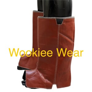 May include: Brown leather chaps with white stitching and the text "Wookiee Wear" in yellow. The chaps are knee-high and appear to be new. The leather has a slight sheen.
